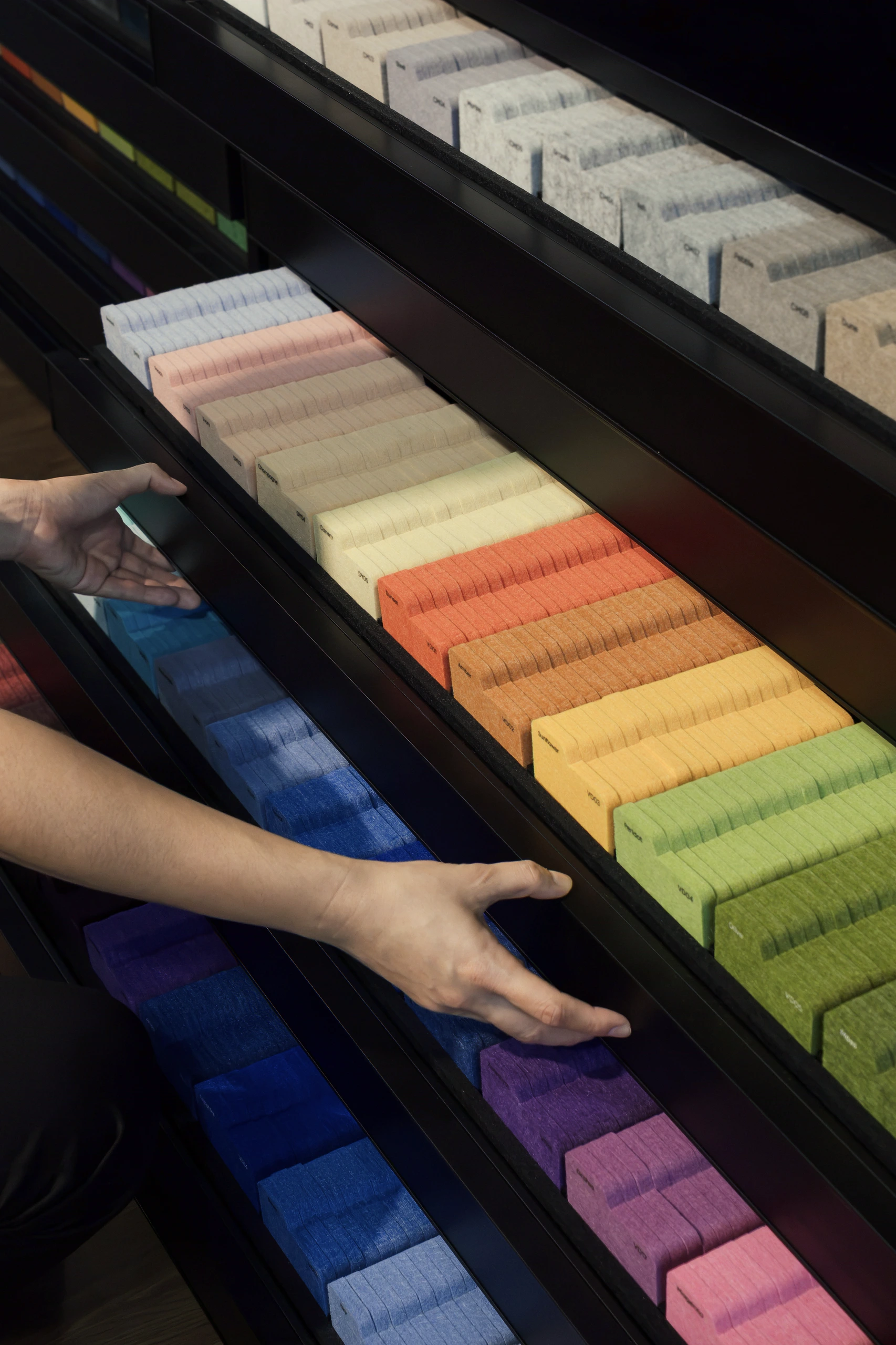 Hands reaching into the Klar™ Material Library, with rows of PET acoustic panel samples arranged by colour